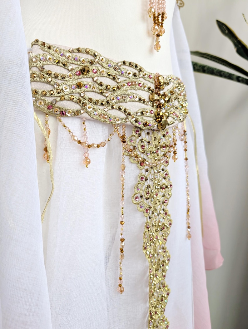 May include: A white garment featuring a wide, ornate gold belt embellished with intricate embroidery, metallic sequins, and small pink and amber beads. A central decorative panel hangs from the belt, adorned with dangling beaded fringe strands.