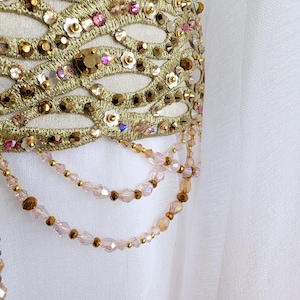 May include: A white fabric garment featuring an ornate gold embroidered lattice overlay. The design is embellished with pink and gold faceted crystals, small white flower-shaped sequins, and dangling strands of light pink and gold beads.