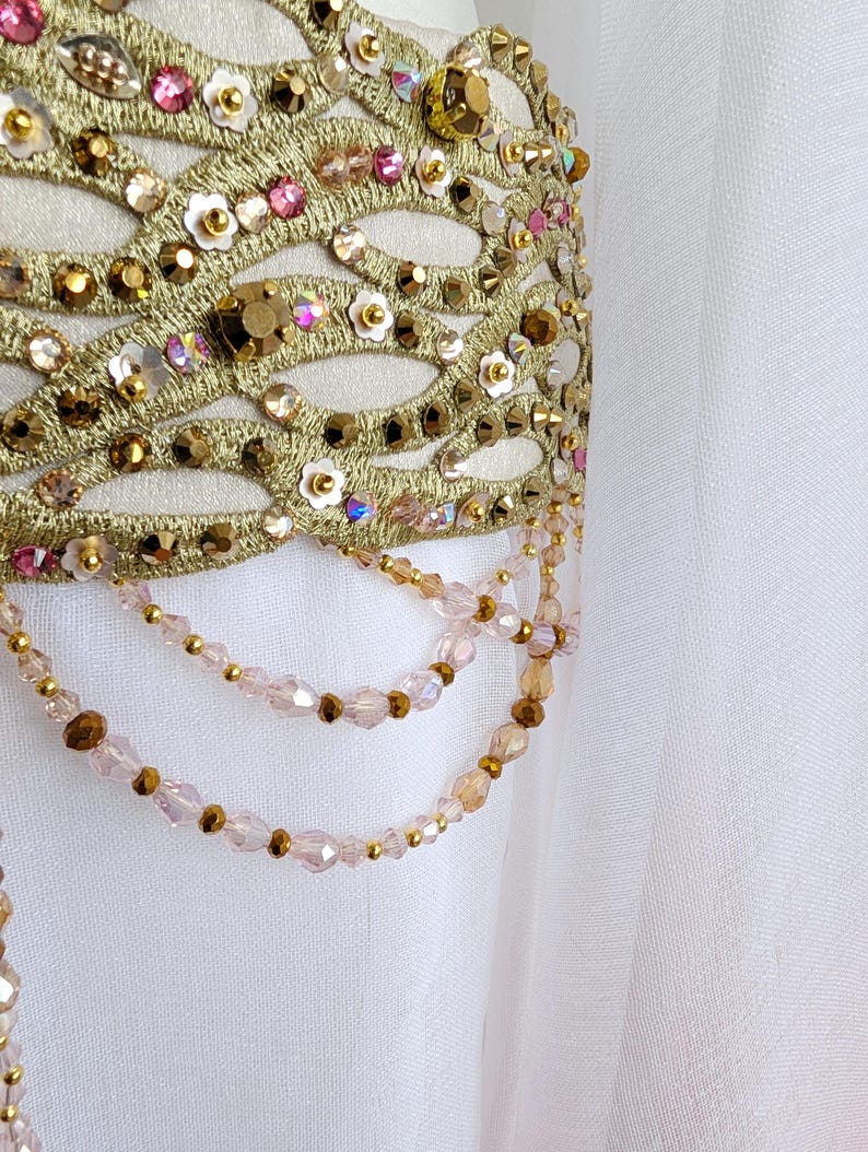 May include: A white fabric garment featuring an ornate gold embroidered lattice overlay. The design is embellished with pink and gold faceted crystals, small white flower-shaped sequins, and dangling strands of light pink and gold beads.