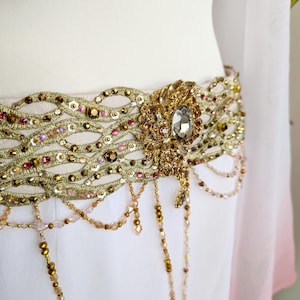 May include: A decorative waist belt featuring an intricate gold-toned lattice pattern embellished with small pink and gold sequins, beads, and crystals. A large central oval crystal ornament serves as a focal point, with dangling beaded fringe.