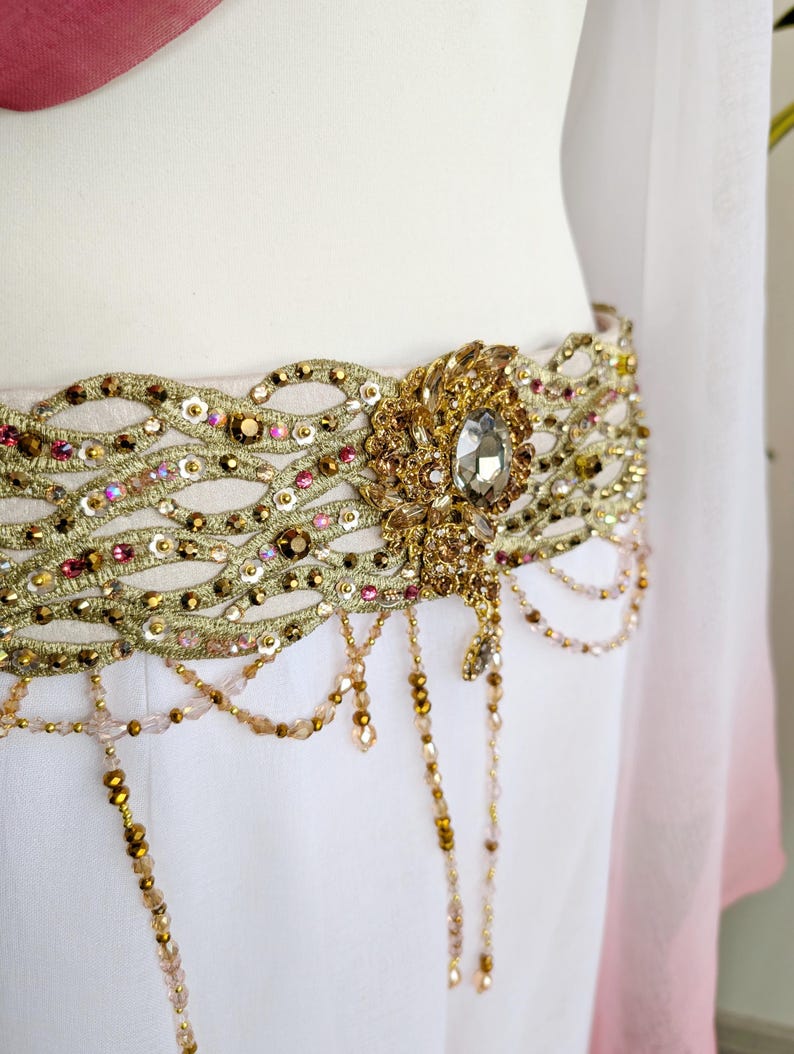May include: A decorative waist belt featuring an intricate gold-toned lattice pattern embellished with small pink and gold sequins, beads, and crystals. A large central oval crystal ornament serves as a focal point, with dangling beaded fringe.