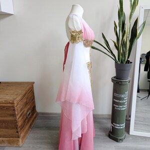 May include: A floor-length dress featuring a white bodice that transitions into a pink ombre skirt. The garment includes long, flowing white-to-pink gradient sleeves, gold-toned embellished shoulder cuffs, and a matching gold-toned waist detail.