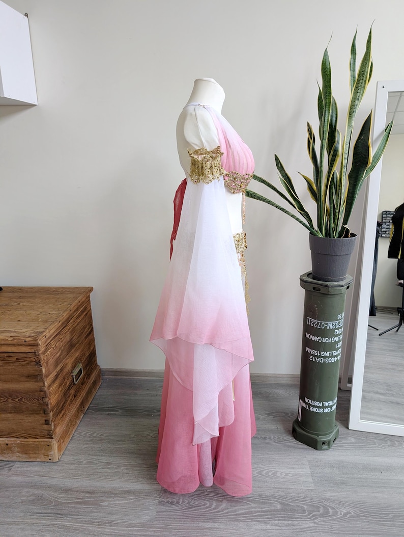 May include: A floor-length dress featuring a white bodice that transitions into a pink ombre skirt. The garment includes long, flowing white-to-pink gradient sleeves, gold-toned embellished shoulder cuffs, and a matching gold-toned waist detail.