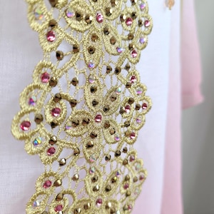 May include: A vertical strip of gold-colored lace embroidery featuring a floral pattern embellished with small, round pink crystals and iridescent, faceted metallic studs. The lace is applied to a smooth, light pink fabric background.