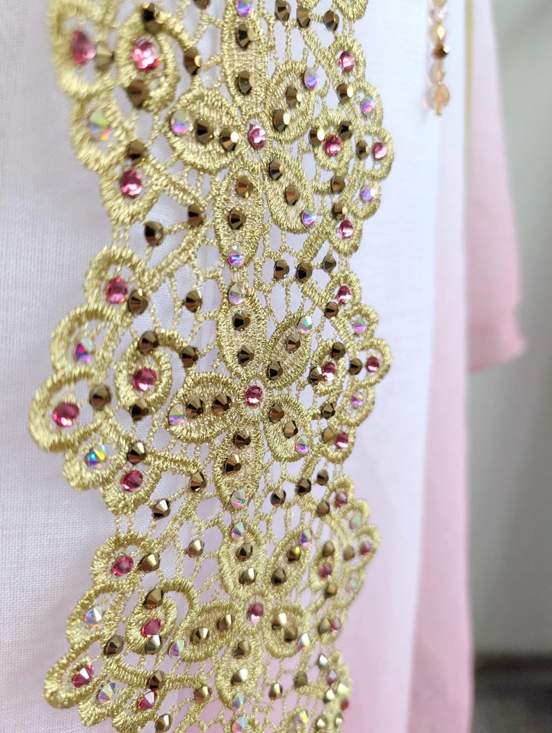 May include: A vertical strip of gold-colored lace embroidery featuring a floral pattern embellished with small, round pink crystals and iridescent, faceted metallic studs. The lace is applied to a smooth, light pink fabric background.