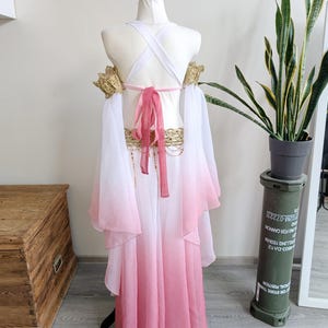 May include: A costume featuring a white and pink ombre skirt and matching flowing sleeves. The bodice includes white crisscross straps, a pink tie closure at the back, and gold-toned decorative lace trim on the shoulders and waistline.