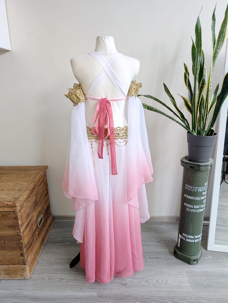 May include: A costume featuring a white and pink ombre skirt and matching flowing sleeves. The bodice includes white crisscross straps, a pink tie closure at the back, and gold-toned decorative lace trim on the shoulders and waistline.