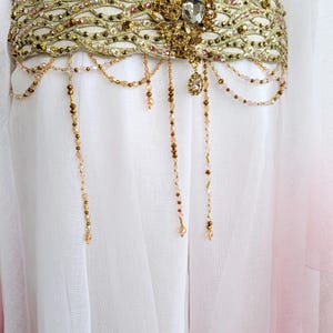 May include: A wide gold belt featuring an intricate woven pattern embellished with small crystals and beads. The belt includes a central ornate gold floral-shaped brooch with a large clear stone and multiple dangling beaded chains in gold and pink.