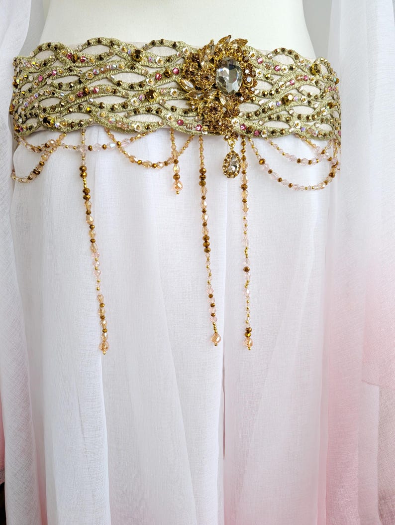 May include: A wide gold belt featuring an intricate woven pattern embellished with small crystals and beads. The belt includes a central ornate gold floral-shaped brooch with a large clear stone and multiple dangling beaded chains in gold and pink.