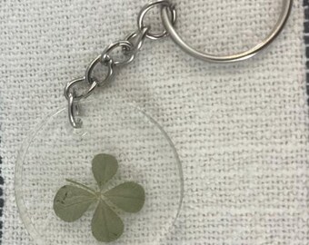 May include: A silver key chain with a clear round charm containing a pressed four-leaf clover. The charm is attached to a silver ring and a silver chain.