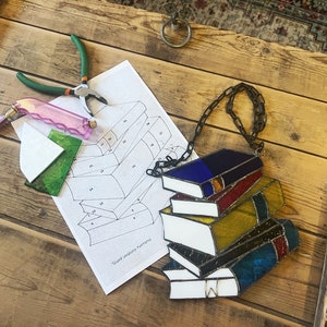 Peut inclure: Une pile de livres en vitrail avec une chaîne noire, avec du verre bleu, rouge, jaune et blanc. Un patron, une pince et des outils sont sur une surface en bois. Les livres sont un objet décoratif.