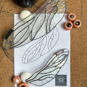 Puede incluir: Dos alas de libélula de vidrio tintado iridiscente con contornos negros. Las alas están numeradas del 1 al 11. Las alas están sobre una hoja de papel blanca con un contorno en blanco y negro de una libélula. Hay pequeñas cuentas de vidrio redondas de varios colores en el papel.