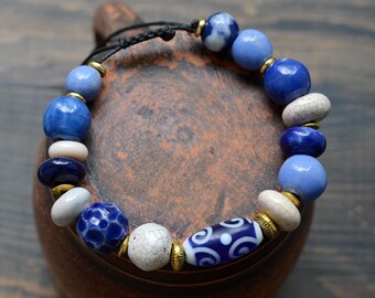 Pode incluir: Um bracelete de contas de cerâmica azul e branco com detalhes dourados. O bracelete é feito de contas de diferentes tamanhos com diferentes padrões e texturas.