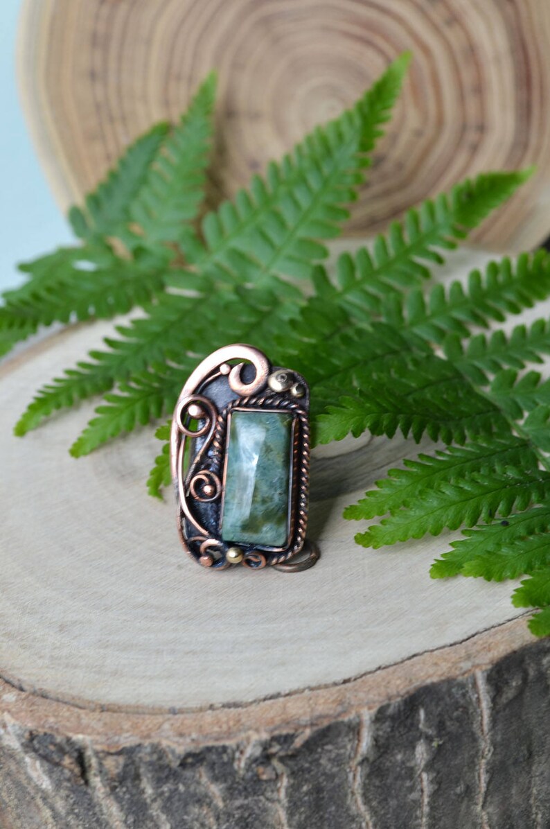K&ouml;nnte beinhalten: Handgefertigter Kupferring mit einem rechteckigen, hellgr&uuml;nen Edelstein. Der Ring weist aufwendige Drahtarbeiten und dekorative Akzente auf. Der Edelstein ist etwa 2 cm lang und 1,3 cm breit. Der Ring ist vor einem Holzgrund mit gr&uuml;nen Farnbl&auml;ttern platziert.