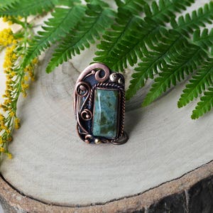 K&ouml;nnte beinhalten: Handgefertigter Kupferring mit einem rechteckigen gr&uuml;nen Stein. Der Ring hat aufwendige Drahtarbeiten und eine dunkle Patina, vor einem h&ouml;lzernen Hintergrund mit Farn und gelben Blumen.