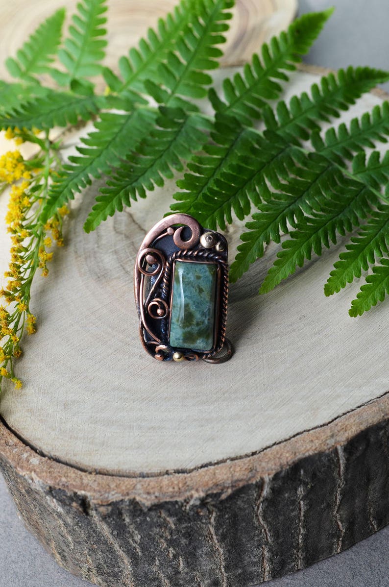 K&ouml;nnte beinhalten: Handgefertigter Kupferring mit einem rechteckigen gr&uuml;nen Stein. Der Ring hat aufwendige Drahtarbeiten und eine dunkle Patina, vor einem h&ouml;lzernen Hintergrund mit Farn und gelben Blumen.