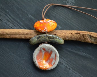 May include: A ceramic necklace featuring three pendants. The top pendant is orange and white, the middle pendant is green and brown, and the bottom pendant is white with orange accents. The necklace is made with a brown leather cord.