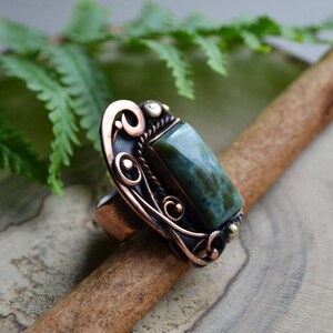 K&ouml;nnte beinhalten: Ein kupferfarbener Ring mit einem l&auml;nglichen, rechteckigen gr&uuml;nen Stein. Der Ring hat verzierte, wirbelnde Filigranmuster um den Stein und auf dem Band. Kleine goldfarbene Akzente sind sichtbar. Der Ring wird auf einer Holzoberfl&auml;che pr&auml;sentiert.