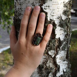 K&ouml;nnte beinhalten: Ein kupferfarbener Ring mit einem rechteckigen, gr&uuml;nen Stein in einem dekorativen Rahmen. Der Ring befindet sich an einer Hand, mit einem Birkenstamm im Hintergrund. Das Design des Rings umfasst verzierte Details und eine dunkle Patina.
