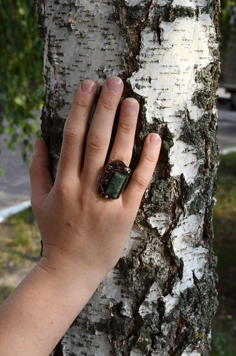 K&ouml;nnte beinhalten: Ein kupferfarbener Ring mit einem rechteckigen, gr&uuml;nen Stein in einem dekorativen Rahmen. Der Ring befindet sich an einer Hand, mit einem Birkenstamm im Hintergrund. Das Design des Rings umfasst verzierte Details und eine dunkle Patina.