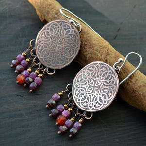 Puede incluir: Un par de pendientes de color cobre con intrincados diseños circulares y colgantes con cuentas en tonos morados, rojos y dorados. Los pendientes se exhiben sobre un palo de madera sobre un fondo oscuro.