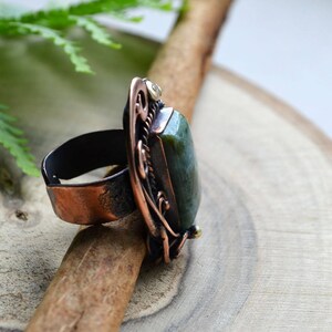 K&ouml;nnte beinhalten: Ein handgefertigter Ring mit einem gro&szlig;en, rechteckigen gr&uuml;nen Stein, der in einen kupferfarbenen Metallrahmen gefasst ist. Der Ring weist aufwendige Filigranarbeiten und ein dunkles Band auf. Der Ring wird auf einer Holzoberfl&auml;che pr&auml;sentiert.