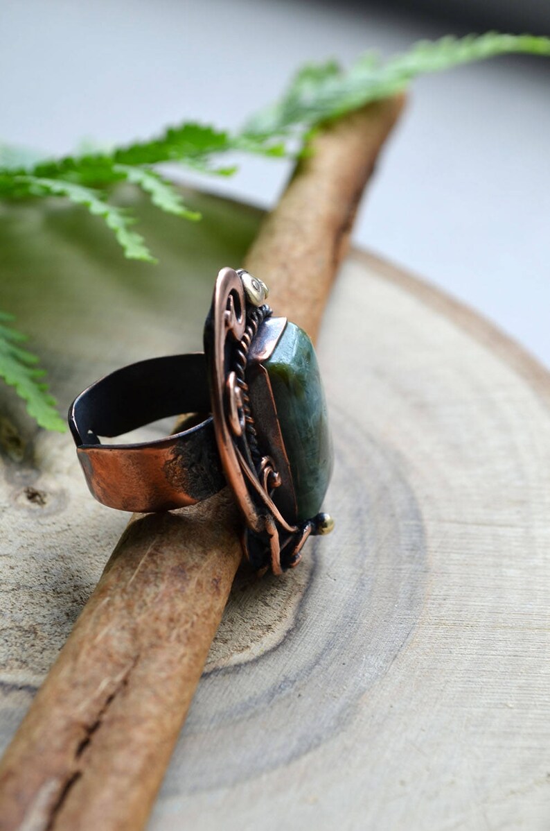K&ouml;nnte beinhalten: Ein handgefertigter Ring mit einem gro&szlig;en, rechteckigen gr&uuml;nen Stein, der in einen kupferfarbenen Metallrahmen gefasst ist. Der Ring weist aufwendige Filigranarbeiten und ein dunkles Band auf. Der Ring wird auf einer Holzoberfl&auml;che pr&auml;sentiert.