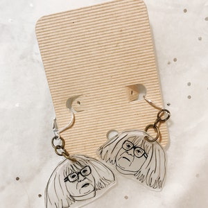 May include: A pair of clear acrylic earrings featuring a black and white line drawing of a woman with glasses. The earrings are hanging from gold-toned hooks.