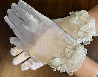 Puede incluir: Un par de guantes de satén blanco con adornos de encaje floral y perlas. Los guantes tienen un diseño clásico, con una textura suave y elegante. Los puños están adornados con delicadas flores de encaje en capas y pequeñas perlas.
