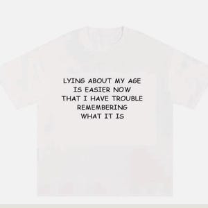 May include: A white short-sleeved t-shirt featuring black text centred on the chest that reads: LYING ABOUT MY AGE IS EASIER NOW THAT I HAVE TROUBLE REMEMBERING WHAT IT IS. The garment has a crew neckline and a standard fit with short sleeves.