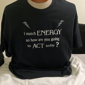 May include: A black short-sleeved t-shirt featuring white embroidered text that reads, I match ENERGY so how are you going to ACT today? The design includes two small grey lightning bolt icons positioned above the text on the chest area.