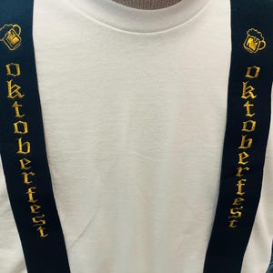May include: Black suspenders with gold embroidered text reading "Oktoberfest". The suspenders feature a gold embroidered beer mug design. The suspenders are worn over a white t-shirt. The text is in a decorative font.