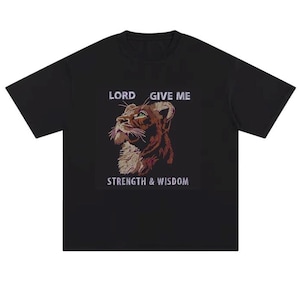 May include: A black short-sleeved t-shirt featuring a central graphic of a lion's head in profile. The text "LORD GIVE ME" appears above the lion, and "STRENGTH & WISDOM" is printed below the image in a light-coloured, sans-serif font.