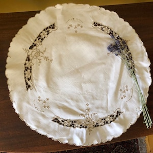 Op de afbeelding: Ronde, witte, geborduurde tafelkleed met een gegolfde rand en delicate bloemmotieven. Een takje lavendel ligt op de tafelkleed.