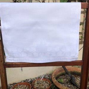 Puede incluir: Una toalla de té de lino blanco con un diseño bordado cuelga de un estante de madera. La toalla está limpia y crujiente, con un brillo sutil.