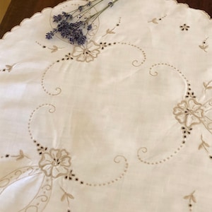 May include: A white round tablecloth with a scalloped edge and delicate brown embroidery of flowers and swirls. A sprig of lavender is placed on top of the tablecloth.
