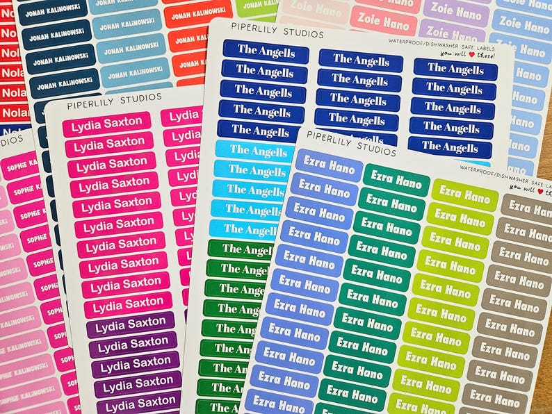 May include: Sheets of personalized name labels in various colors, including pink, blue, green, and purple. The labels feature names like "Lydia Saxton," "The Angells," and "Ezra Hano" in white text. The labels are rectangular and designed for various uses.