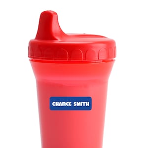 May include: A red sippy cup with a matching lid. A blue rectangular label on the cup reads "CHANCE SMITH" in white capital letters. The cup is designed for toddlers and young children.