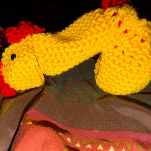 May include: A handmade, yellow crocheted rooster with red accents on the comb and wattle. The rooster has a black eye and is lying on a patterned fabric. The craft item is a whimsical decorative piece.