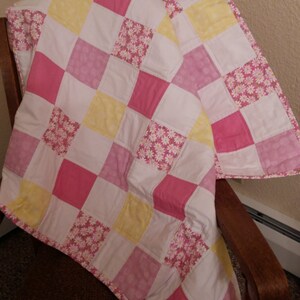 May include: A patchwork quilt with a checkered pattern in pink, yellow, and white. The quilt is made of squares of different fabrics, some with a floral print. The quilt is folded and draped over a wooden chair.