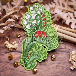 May include: Embroidered patch featuring a stylized floral design with white flowers, red berries, and green leaves. The patch has a light green border and is set against a wooden background with decorative elements.