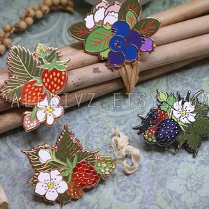 May include: Enamel pins featuring colorful berry designs, including strawberries, blueberries, and blackberries, with gold-colored outlines. The pins are arranged on wooden dowels and a patterned surface.