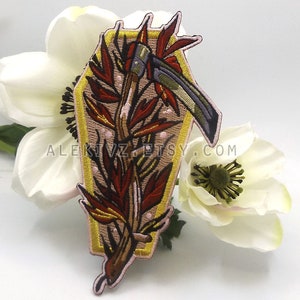 May include: Embroidered patch featuring a coffin shape with a scythe and floral elements. The patch has a tan background with a gold border and is decorated with red and brown leaves. The text "ALEKIVZ" is visible.