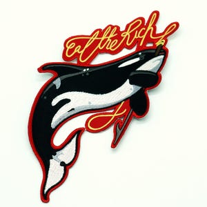 May include: Embroidered patch featuring a black and white orca with a red border. The patch has the text "Eat the Rich" in a stylized, cursive font. The orca is depicted with a defiant expression, adding a touch of rebellion.