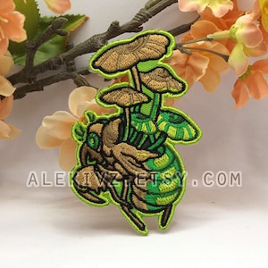 May include: Embroidered patch with a whimsical design of mushrooms on a stylized insect. The patch has brown, green, and yellow colours with a neon green border. The text "ALEK1VZ ETSY.COM" is visible.