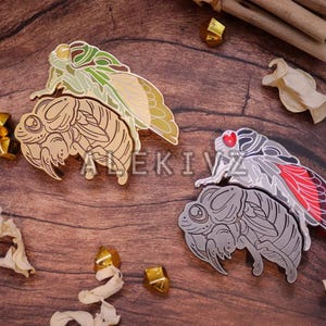 May include: Two cicada-shaped enamel pins with detailed wing designs. One pin features green and gold wings, the other has red and gray wings. The pins are on a wooden surface with decorative elements.