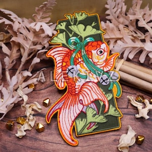 May include: Embroidered patch featuring a vibrant orange and white goldfish with detailed fins and a green accent. The patch includes small silver bells and is set against a green backdrop with leaf designs. Decorative item.