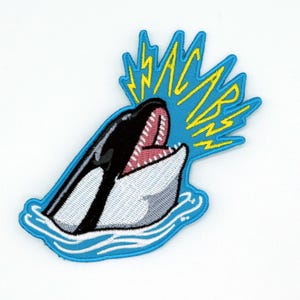 May include: Embroidered patch featuring a stylized orca breaching the water. The orca is black and white with a pink mouth. Above the orca is a blue lightning bolt design with the word "WHACARB" in yellow.