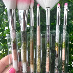 May include: Set of six makeup brushes with clear handles filled with various crystals and herbs. The brushes have pink and white bristles and are arranged in a plastic holder.