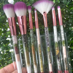 May include: Set of six makeup brushes with clear handles filled with crystals and herbs. The brushes have pink and purple bristles and rose gold colored metal accents.